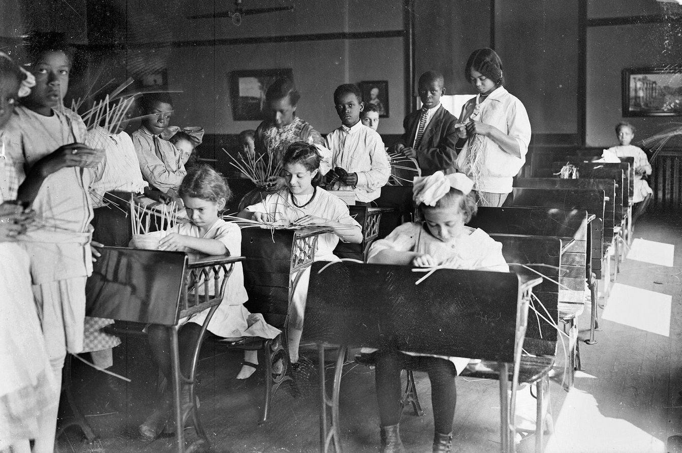 School children, including several African Americans, at Raymond School sitting and standing at their desks and learning basket weaving in the Douglas community area, Chicago, Illinois, 1910s.