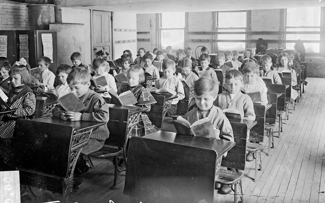 Children reading at desks in a classroom at the Robert Emmet School, looking at the children from the front of the classroom in the Austin community area, Chicago, Illinois, 1910s.