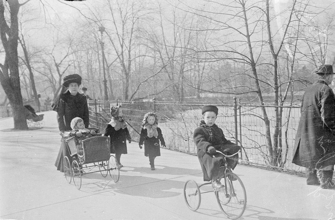 A woman pushing a child in a stroller, with two children walking to the side and a child riding a tricycle in front of them in Lincoln Park in spring, Chicago, Illinois, 1910s.