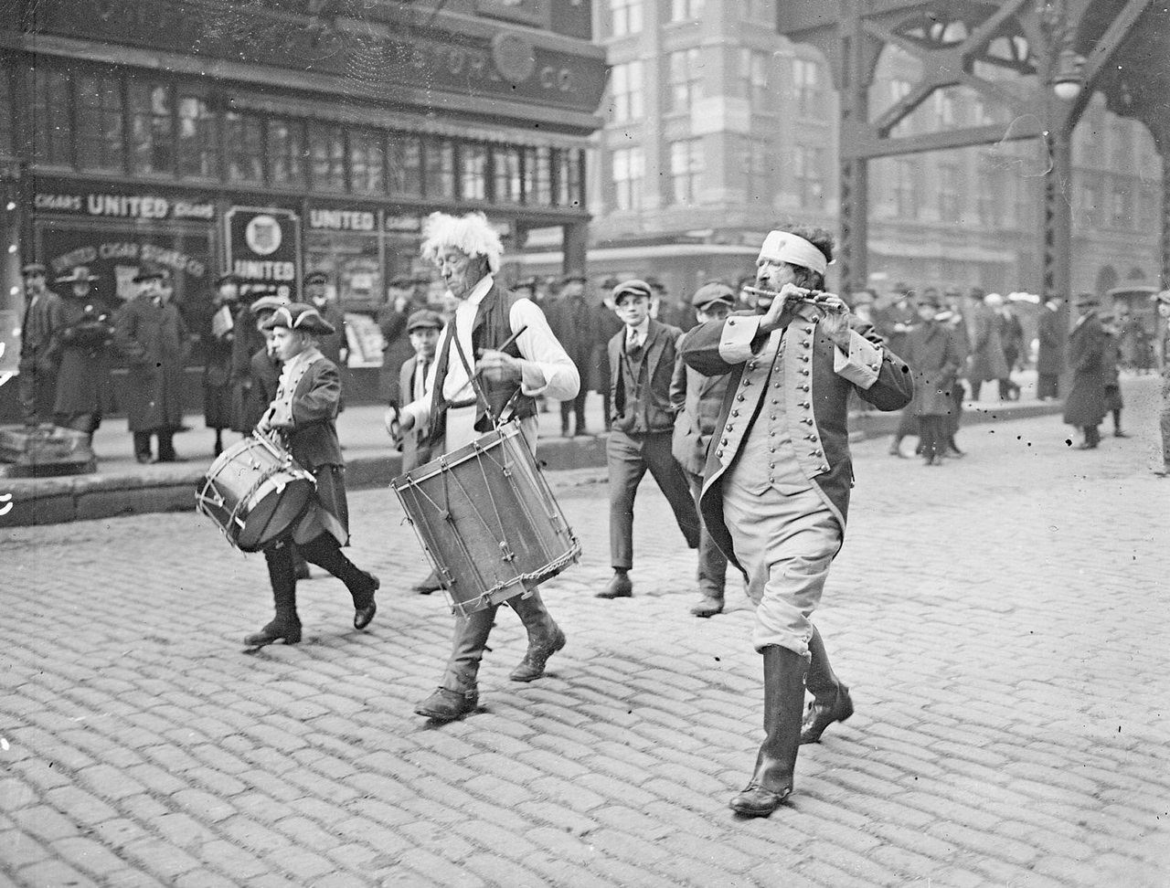 Men dressed in Colonial costumes, marching south and playing drums and a fife on Market Street in the Loop community area, Chicago, Illinois, 1910s.