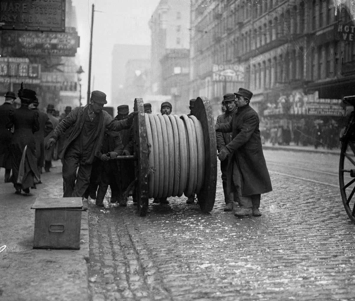 Men rolling a large wheel of underground telephone cable on a business street, Chicago, Illinois, 1910s.