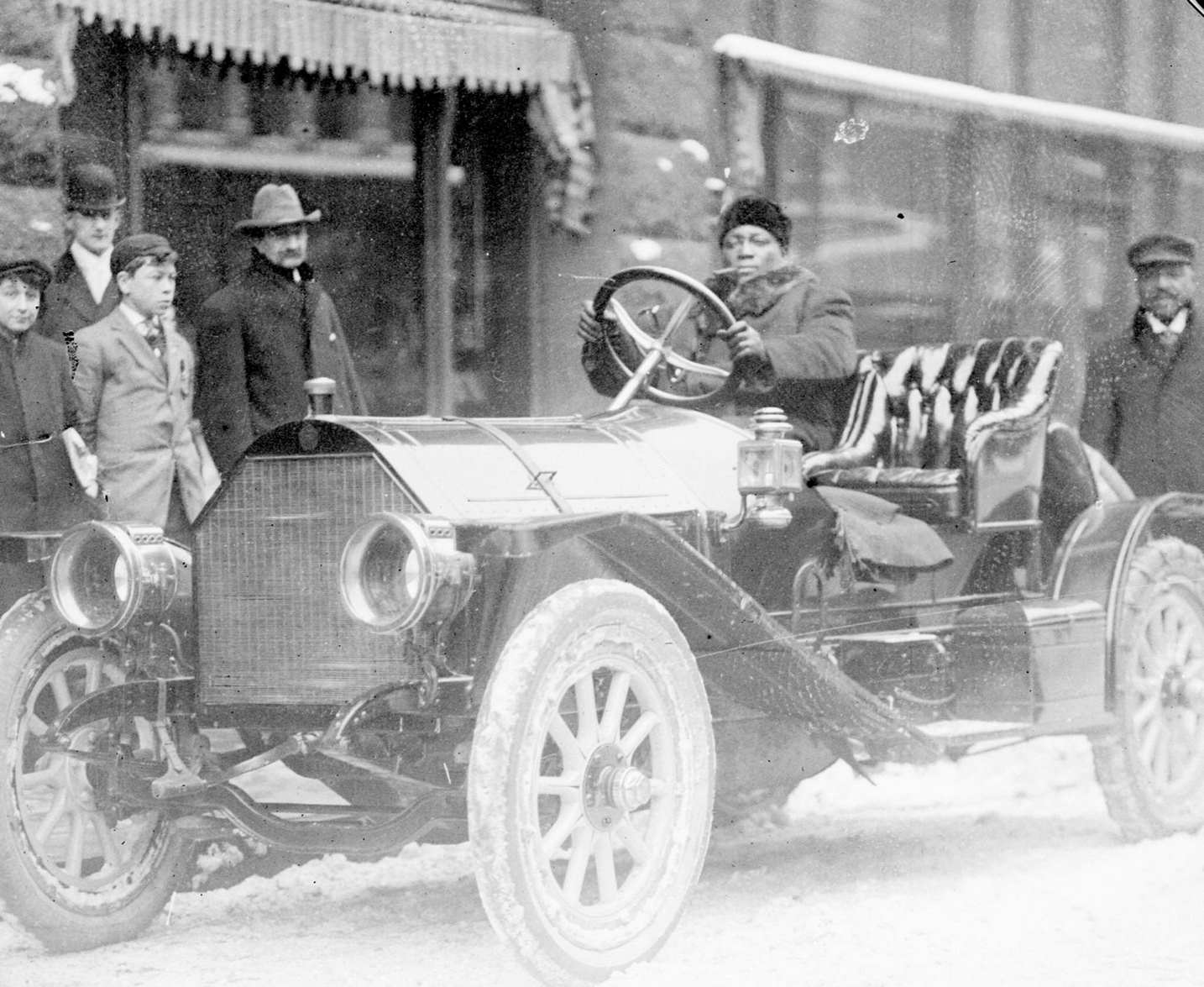 Pugilist Jack Johnson sitting in the driver's seat of a convertible automobile and holding on to the steering wheel on a snow-covered street in Chicago, Illinois, 1910s.