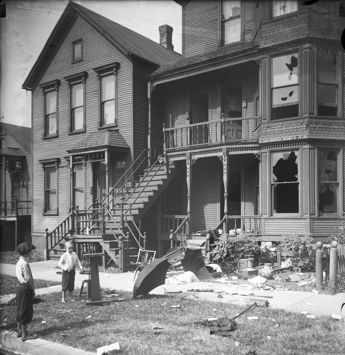 Two young boys outside a home, with broken windows and debris in the front yard, which had been vandalized during a race riot, Chicago, Illinois, July or August 1919.