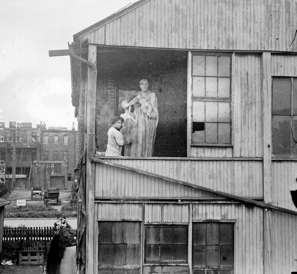 Ida McClelland Stout, an African American sculptor, standing on a tenement porch, next to a monumental Ethiopian sculpture, Chicago, Illinois, 1910s.