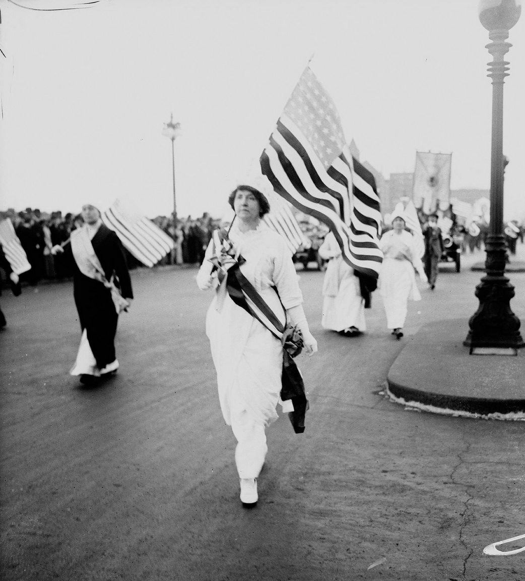 Grace Wilbur Trout wearing a sash, holding an American flag, marching north on South Michigan Avenue, with other suffragists holding flags marching nearby in a women's suffrage parade in the Loop community area of Chicago, Illinois, 1910s
