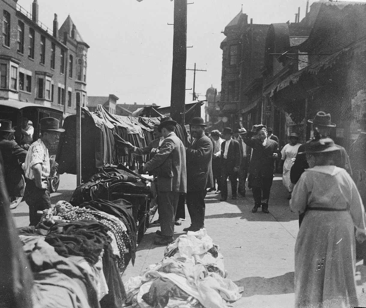 Merchants selling clothing and a group of men inspecting the goods on Maxwell Street at the Sunday market in the city's Jewish community located in the Near West Side community area, Chicago, Illinois, 1917.
