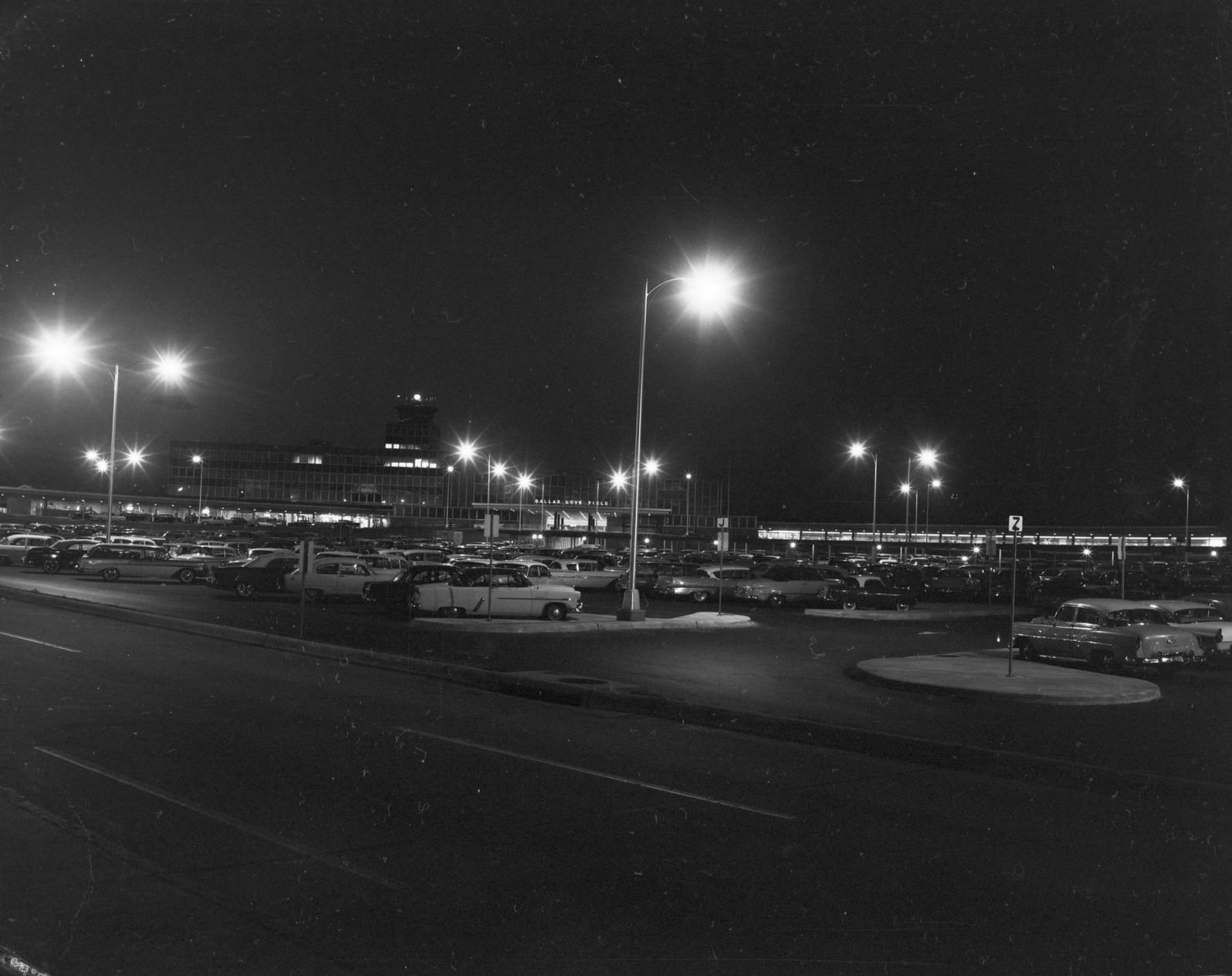 Dallas Love Field at night, 1950. There are lampposts with the lights on. There are automobiles parked on the parking lot.
