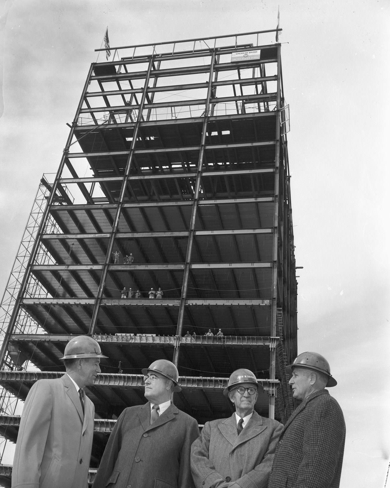 Building under construction, downtown Dallas, 1950 There are four men standing next to each other. Each of them are wearing a safety helmet.