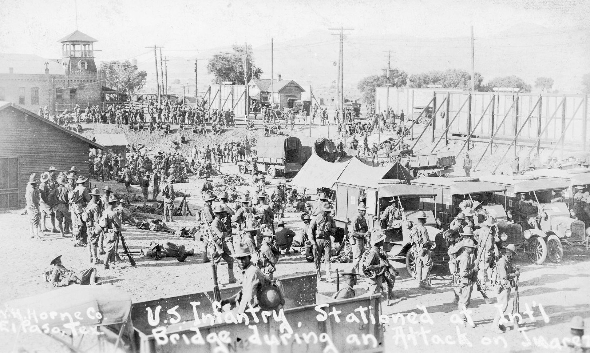 Group shot of many African American members of the United States Infantry, outside and among tents, buildings, and equipment, at the International Bridge in El Paso, 1900s