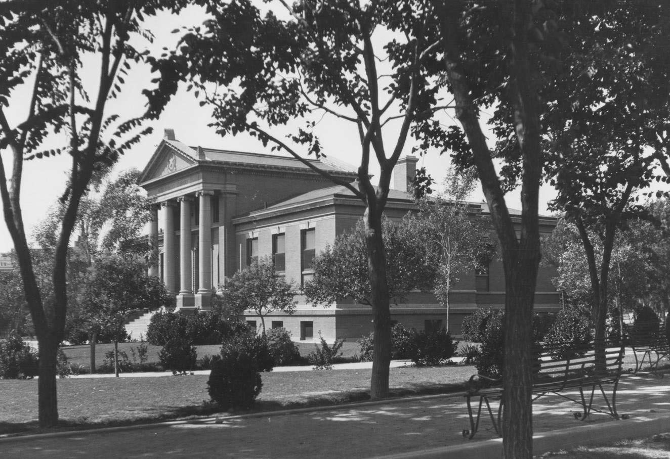 The front and right side of the Carnegie Library in El Paso, 1904. The building has four large ionic columns with a star engraving above them. Trees and bushes can be seen on the landscaping.