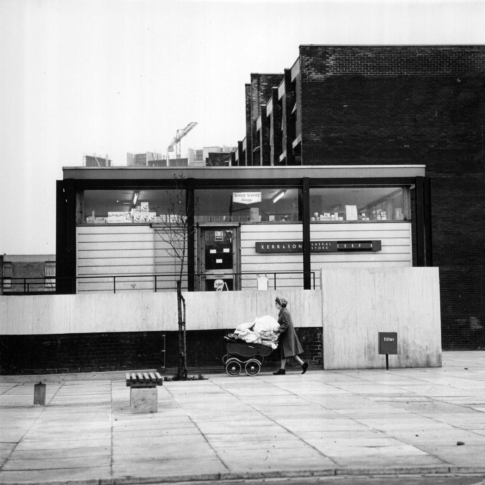 A woman pushing a pram laden with bedding past a newly built shop in the modern Gorbals area of Glasgow, 1960