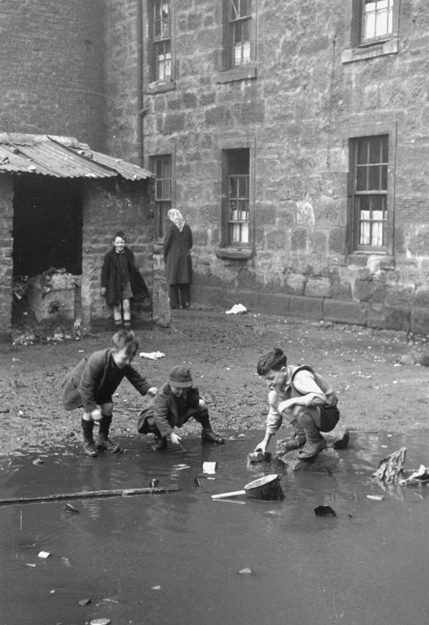 Children playing in a puddle in the Gorbals, a slum distict of Glasgow with graffiti on the walls and muddy pools and rubbish littering the back yards, 1948