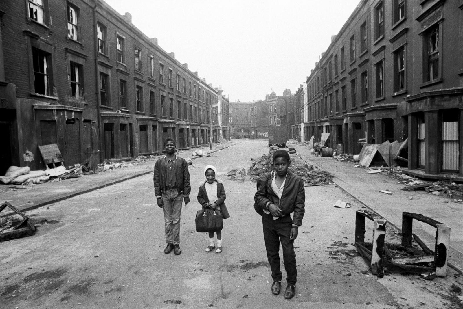 Three children, including a little girl carrying a satchel, walking along a street of slum housing in the notorious Gorbals district of Glasgow in 1969.