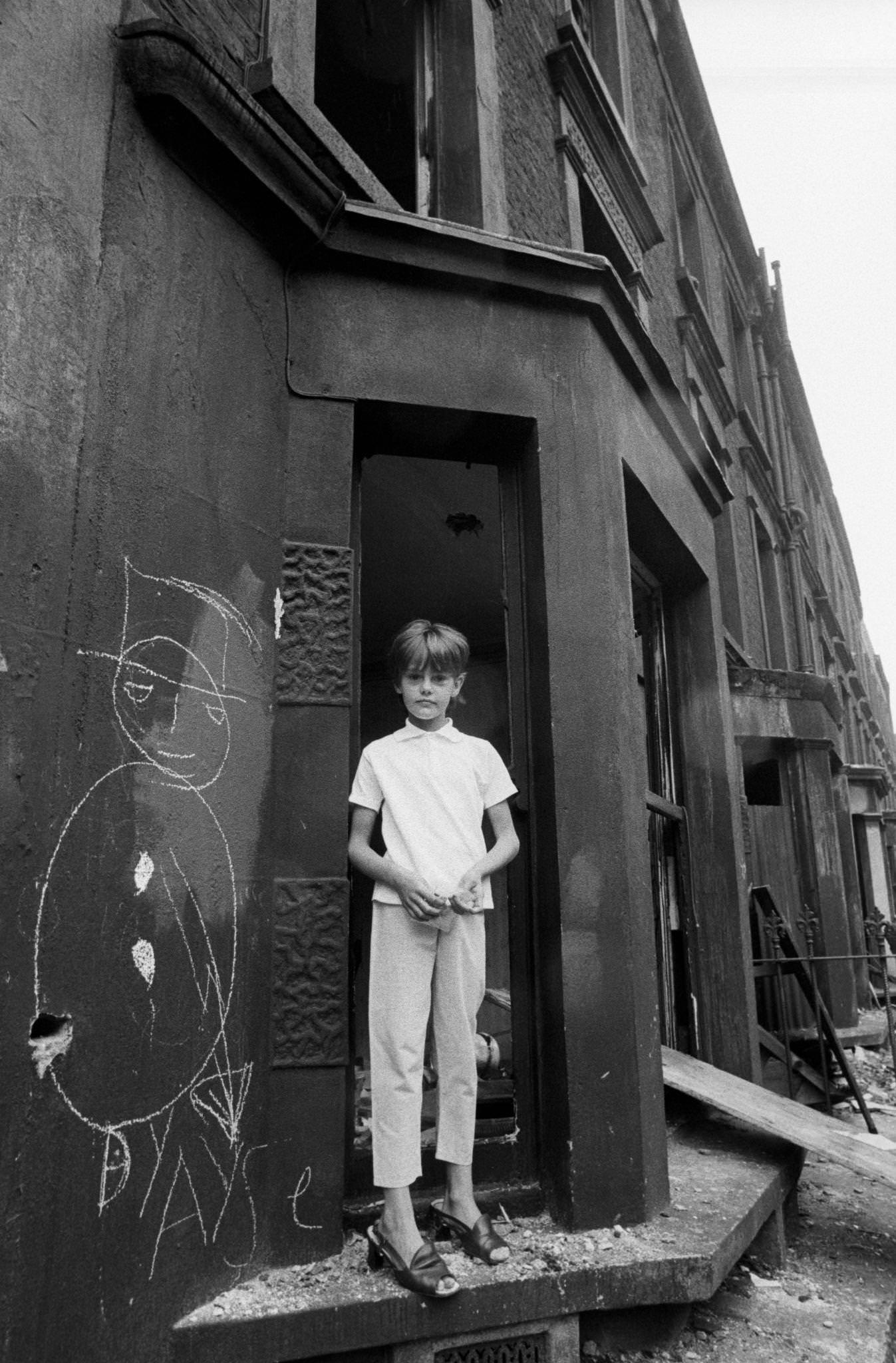 A thin child wearing women's shoes standing in a doorway next to a graffiti figure in slum housing in the notorious Gorbals district of Glasgow in 1969.