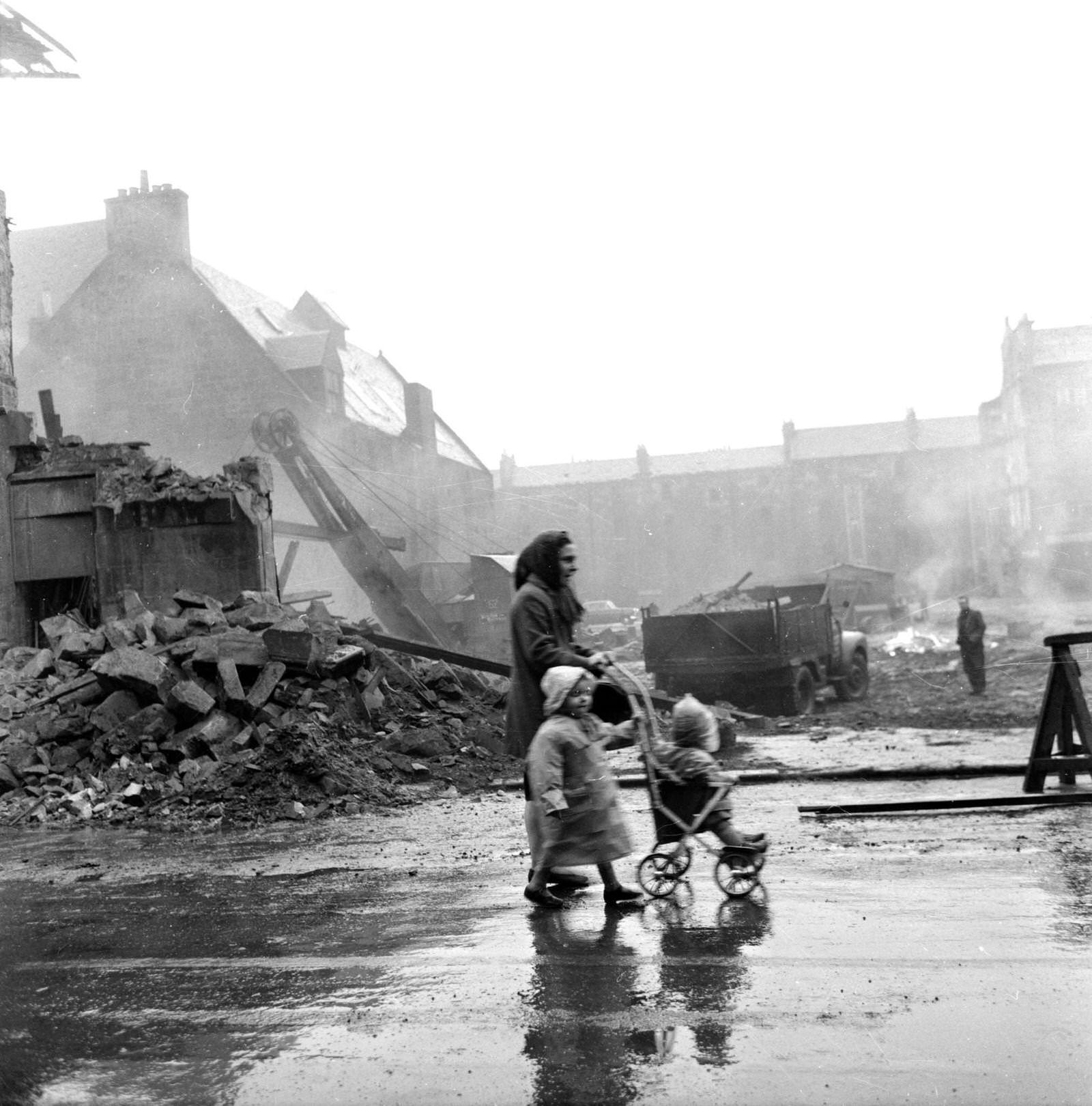 A woman with two young children walking past a demolition site in the Gorbals area of Glasgow, 1962