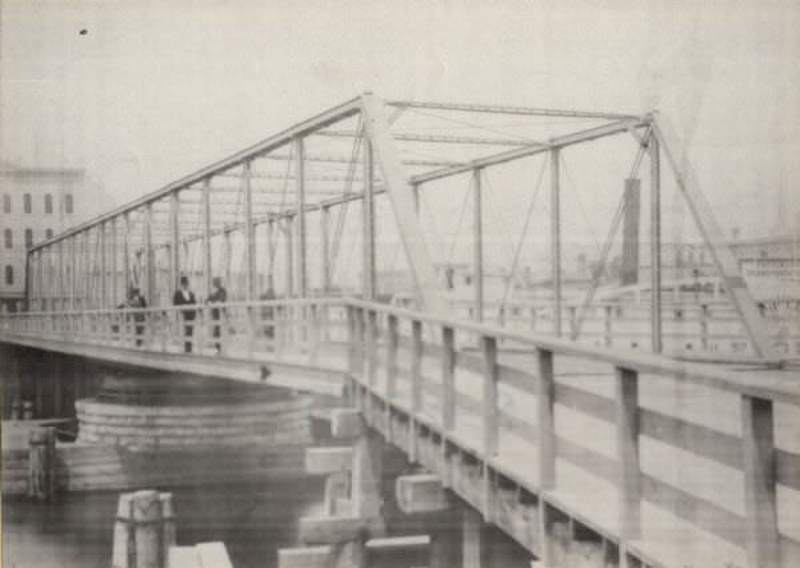 Metal bridge over river with three men standing near the railing, 1870