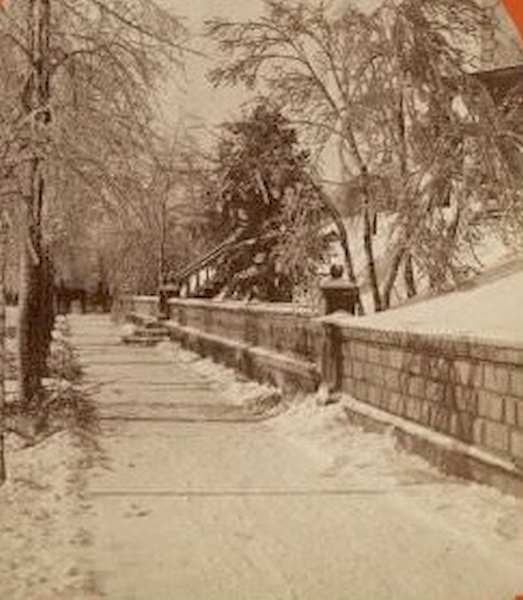 Winter scene of a residential area, looking down a snowy sidewalk near a retaining wall and a fence in Milwaukee, with a horse-drawn carriage or sleigh in the distance, 1876