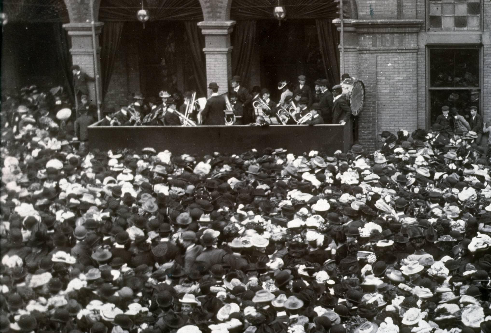 A brass band plays in a boxed-off area outdoors, while crowds gather around at a Sängerfest competition, 1886