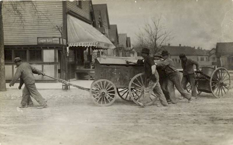 Men pushing and pulling two carts, one with a barrel, around a street corner 1897