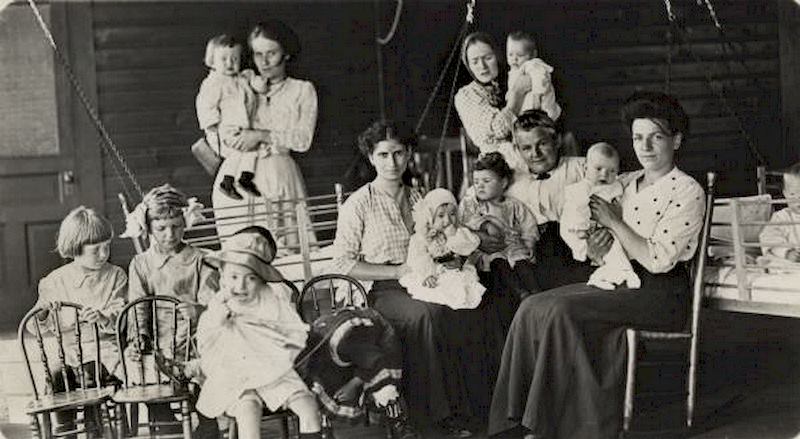 Women of various ages are surrounded by babies and young children, as well as chairs and cribs, 1897