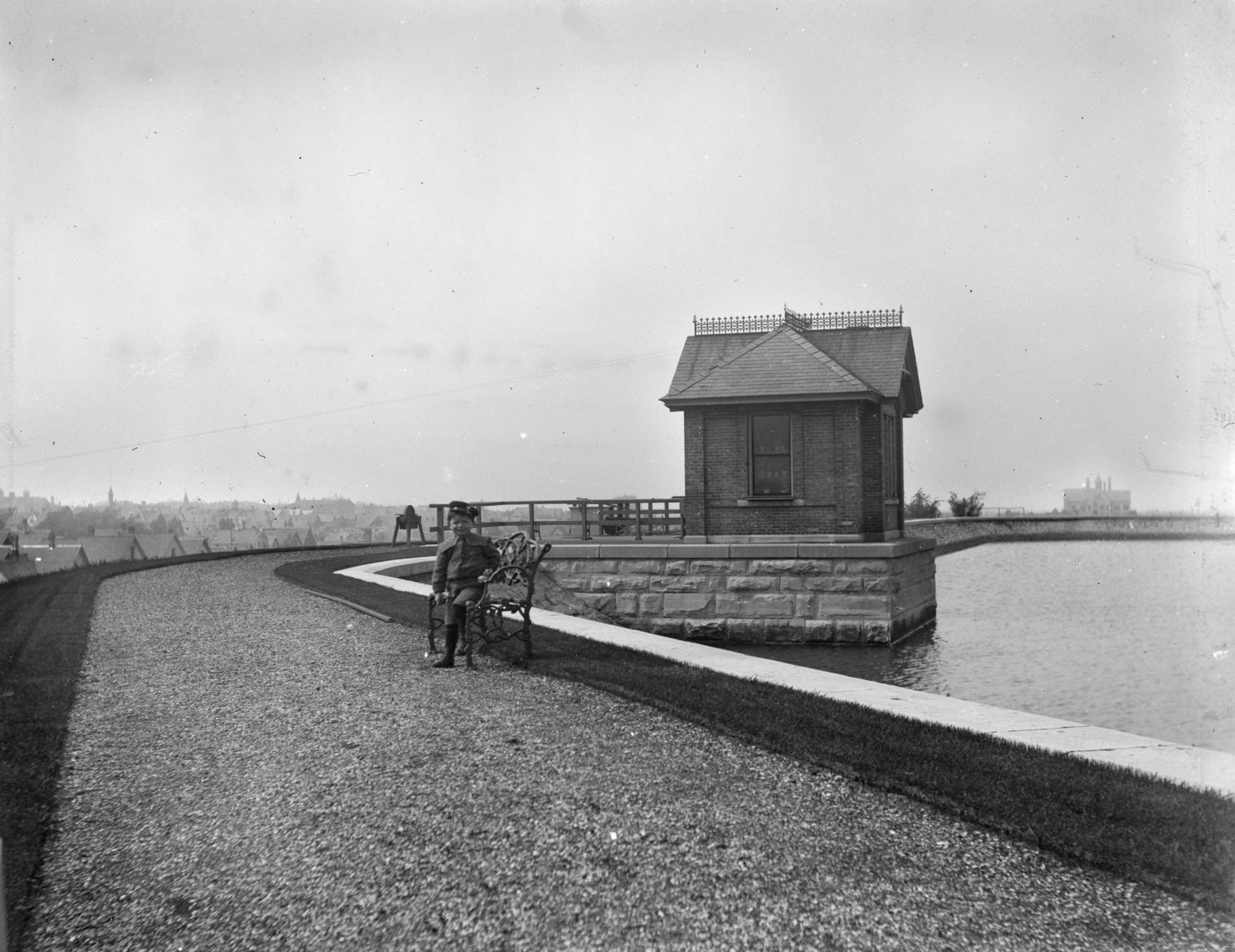 View down gravel path towards Syl seated on a bench along the edge of a reservoir on a hill, Milwaukee, Wisconsin, May 28, 1899.