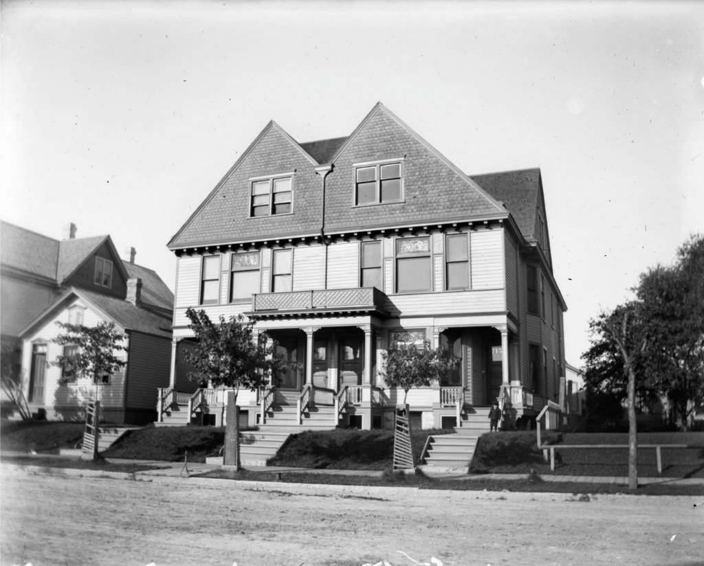 View across road of Syl standing in front of a three-story duplex house on Jones Island, Milwaukee, Wisconsin, 1898.