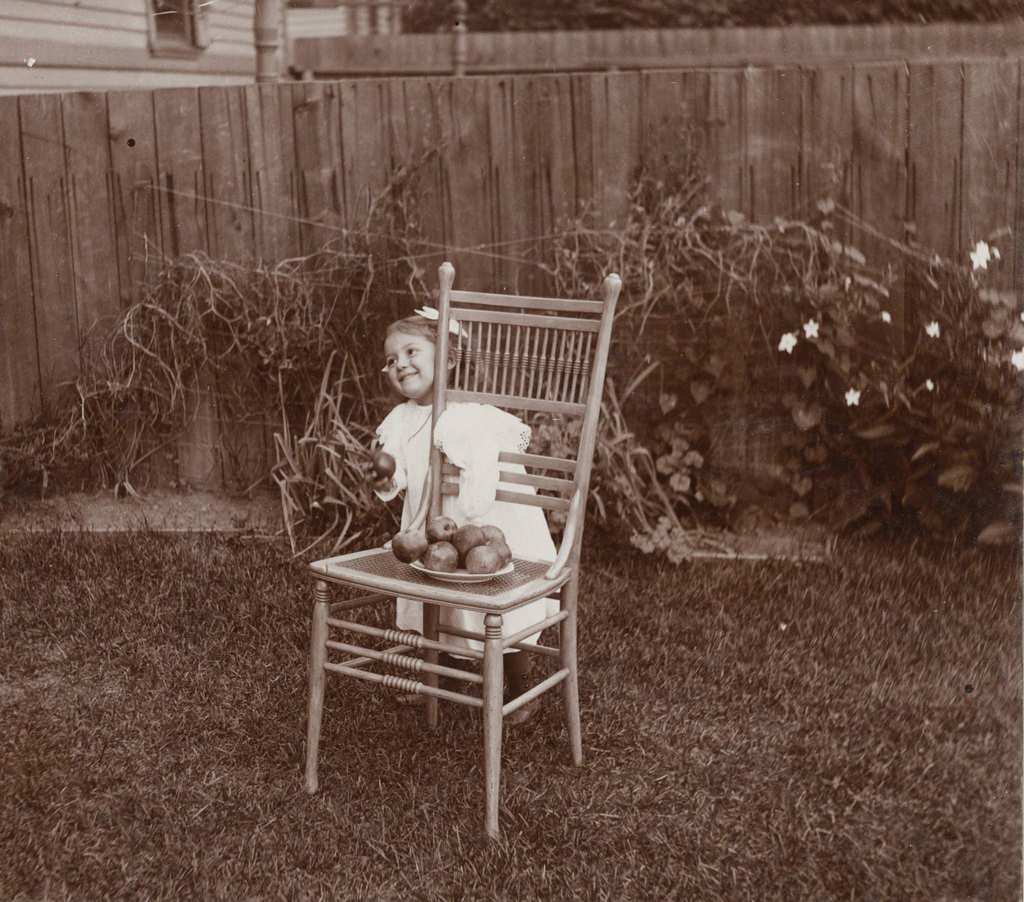 A young girl, Helen Phillips, stands in a yard near a fence behind a wooden chair, Milwaukee, Wisconsin, 1898.