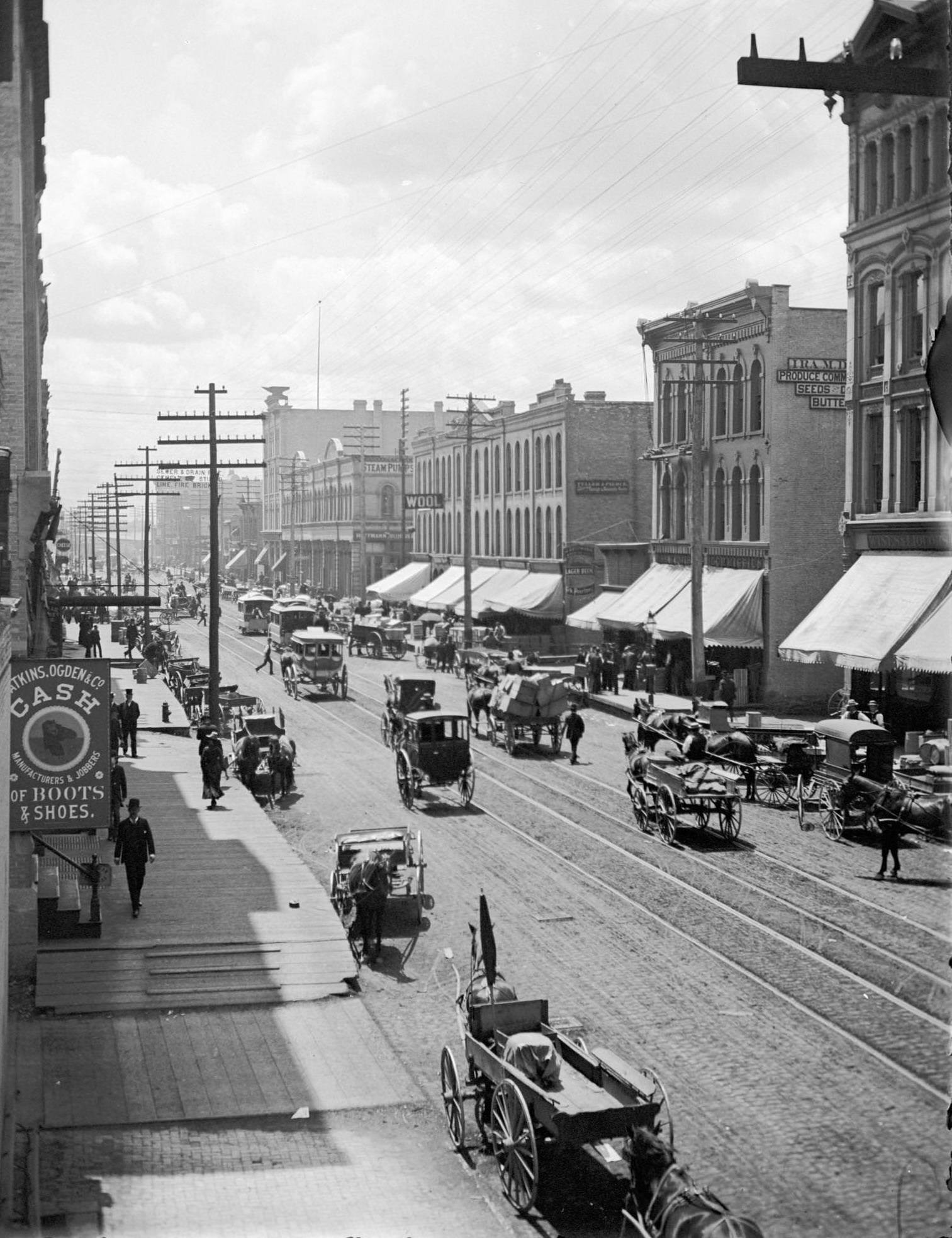 Elevated view of West Water Street from Grand Avenue of horse-drawn vehicles, pedestrians, buildings, storefronts and power lines, Milwaukee, Wisconsin, 1885.