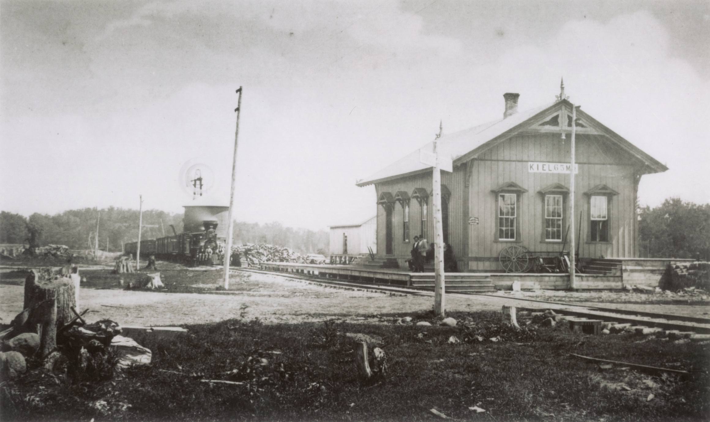 Two men are standing on the platform outside of Kiel’s first Depot, 1870