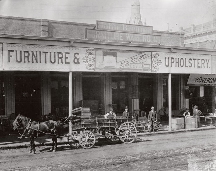 View of the one-story, brick warehouse; horse-drawn Breuner wagon in front; several men standing along wooden sidewalk; goods and boxes stacked on sidewalk, 1882