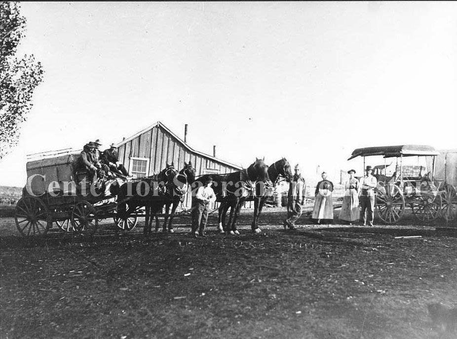 Two stage coaches, one hitched with four horses, one parked and two women and three men standing next to the coaches, 1881