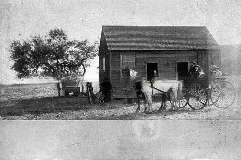 Small country house in middle of field with two small trees, men stand to side, horses and buggy with six people aboard parked in front, 1880