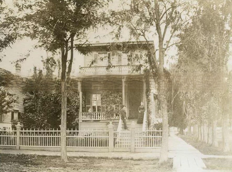 View of two story wood sided home. A man and a woman are on the stairs, 1880s