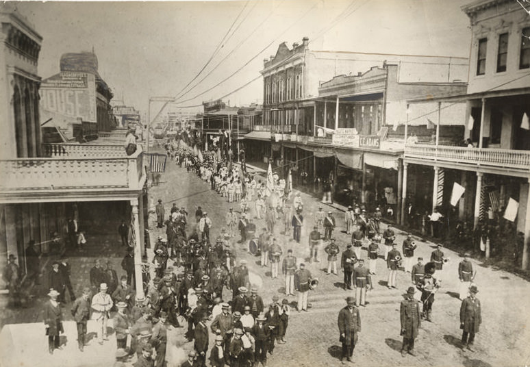 Parade of Turn-Verein group, "Elites Turn-Fest", on J Street, 700 block, looking east. Led by band including bandleader in shako hat. Group met in Sacramento on June 21-23, 1885.