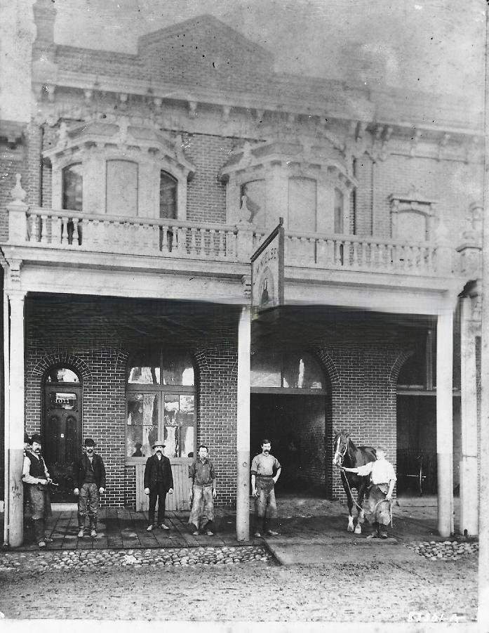 Seven men stand on porch of a stables, one holds reins of horse, 1881