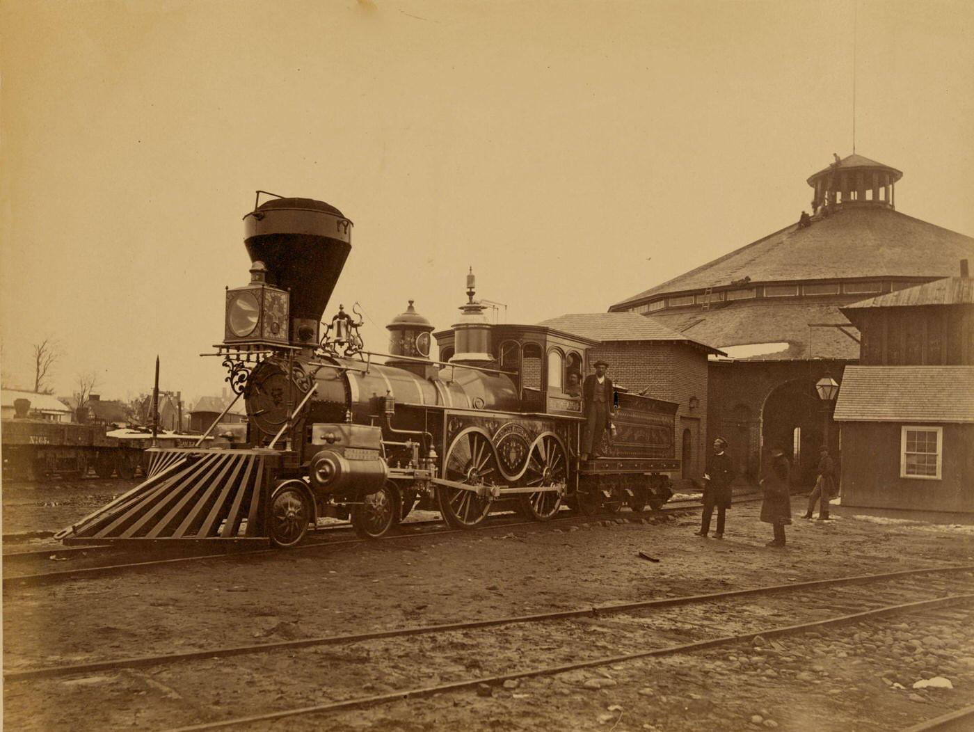 Ornately decorated locomotive J.H. Devereux, of the United States Military Railroad with two crew members on board outside the roundhouse at the Alexandria station, 1863