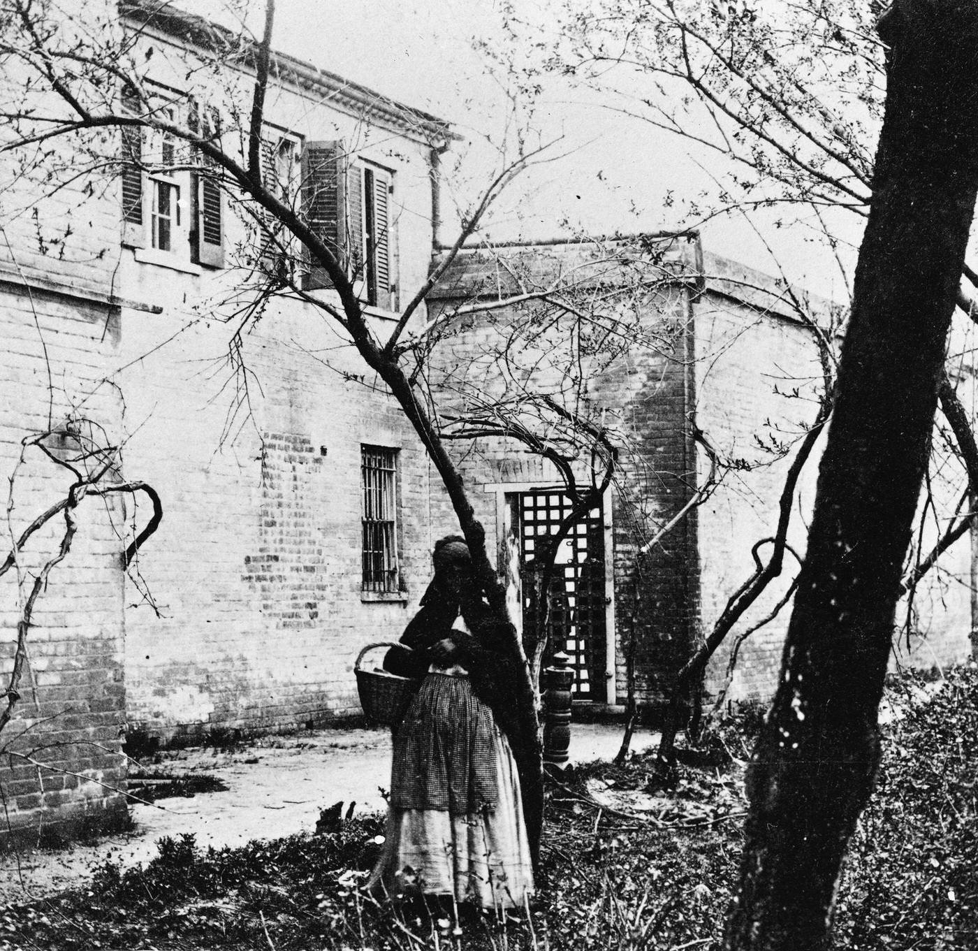 A black woman holds a basket and stands among bare trees in a slave pen, Alexandria, Virginia, 1860s