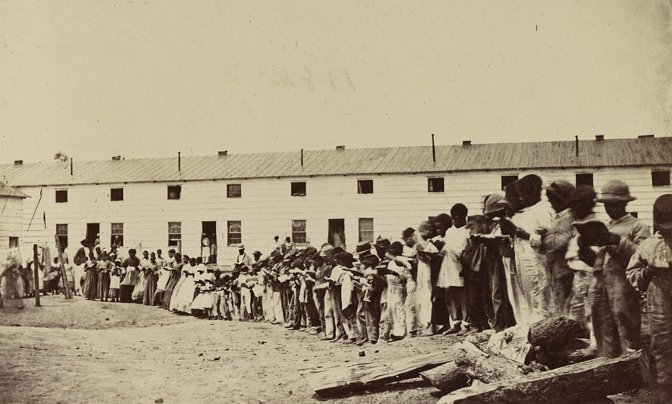 African-American adults and children reading books while lined up in front of a barracks-like building. The same building is shown in "Freedman's Barracks, Alexandria, 1860s
