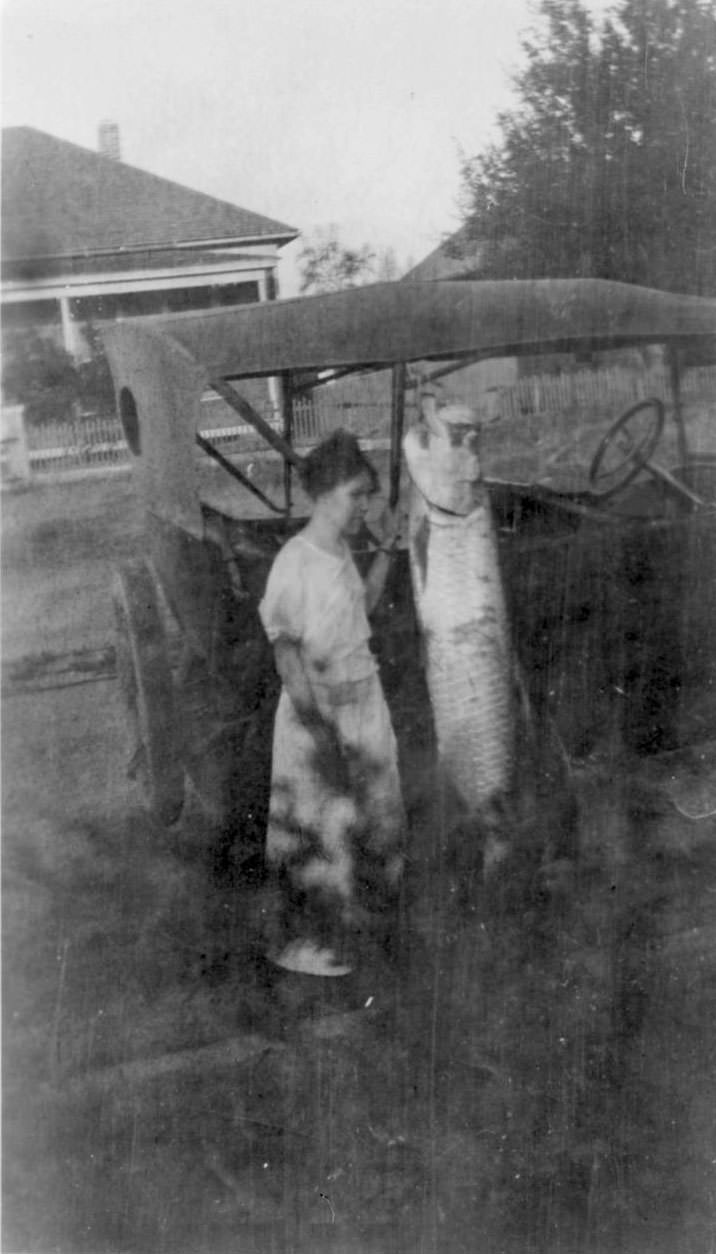 A woman standing next to an automobile. There is a large fish hanging from the roof of the car. There is a house in the background.