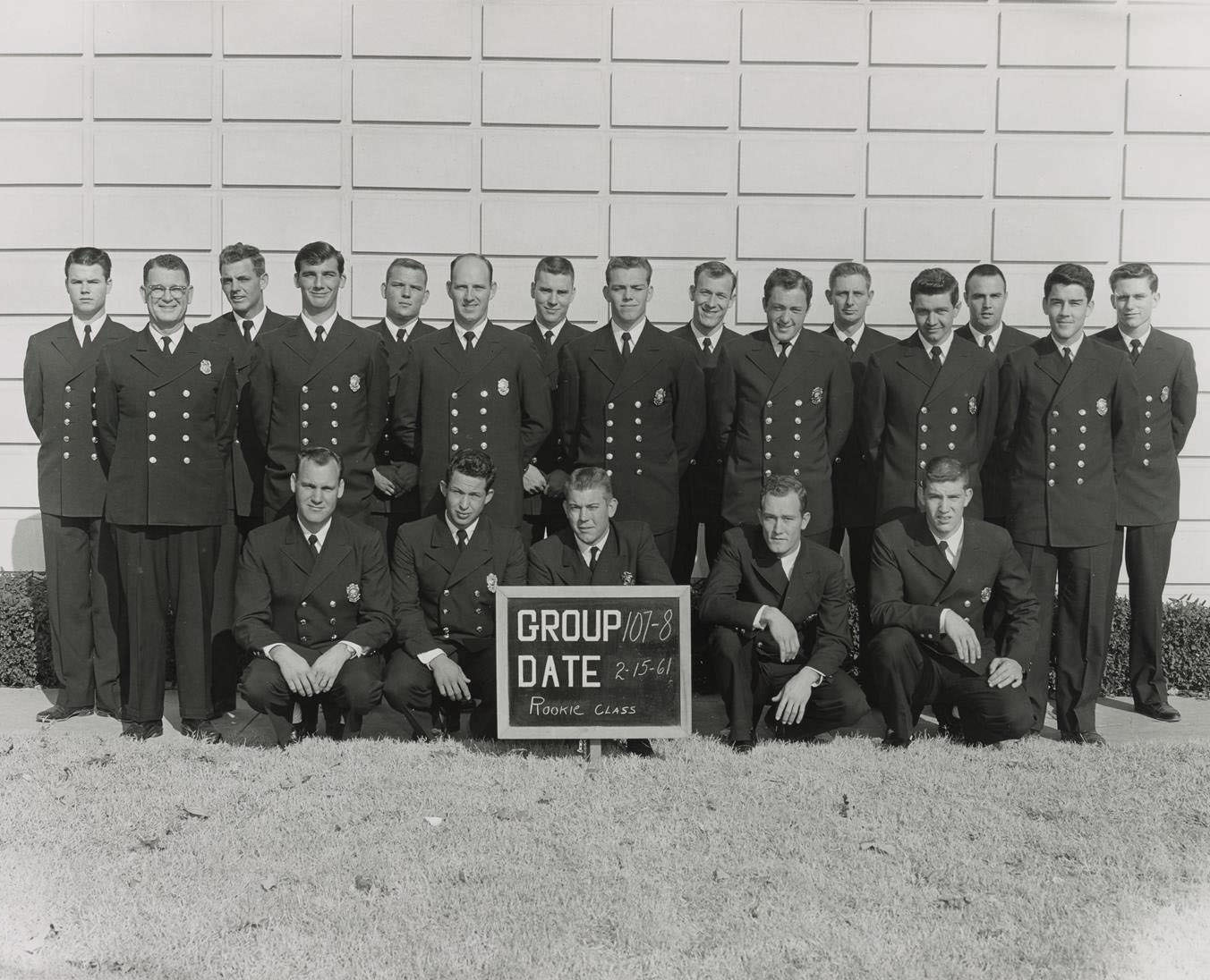 Dallas Fire recruits from class numbers 107 and 108, posing in three lines, standing and kneeling in front of a concrete edifice with a grid pattern, 1967.