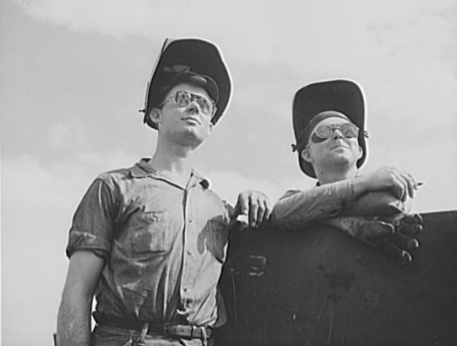 Their helmets hoisted on their heads, these two welders take time out for a smoke and breath of fresh air. Hundreds of welders are employed in the building of Uncle Sam's new Navy craft, 1941