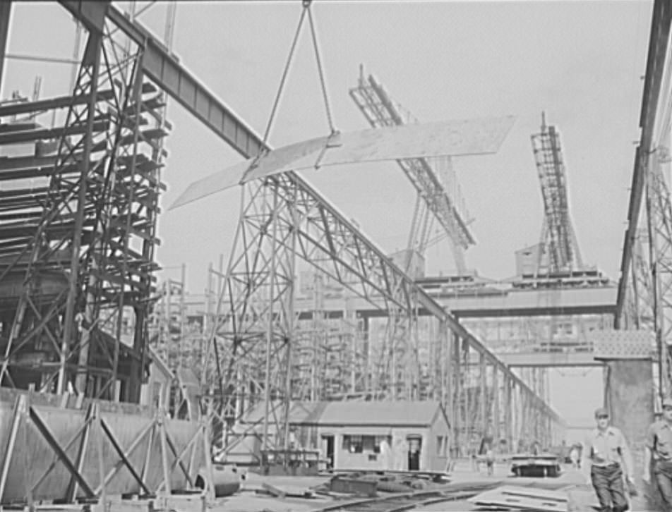 A huge overhead crane swings this metal plate into position to be welded to a new cruiser under construction for the U.S. Navy, 1941