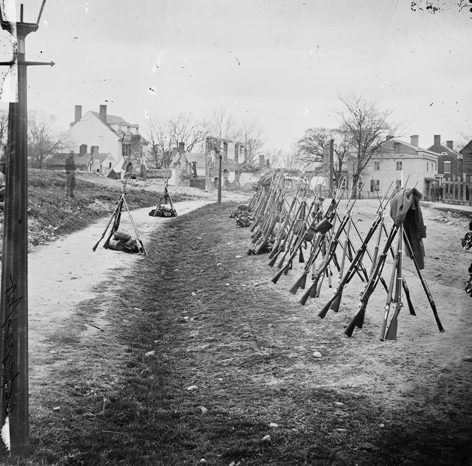 Row of stacked Federal rifles; houses beyond, Petersburg, 1865