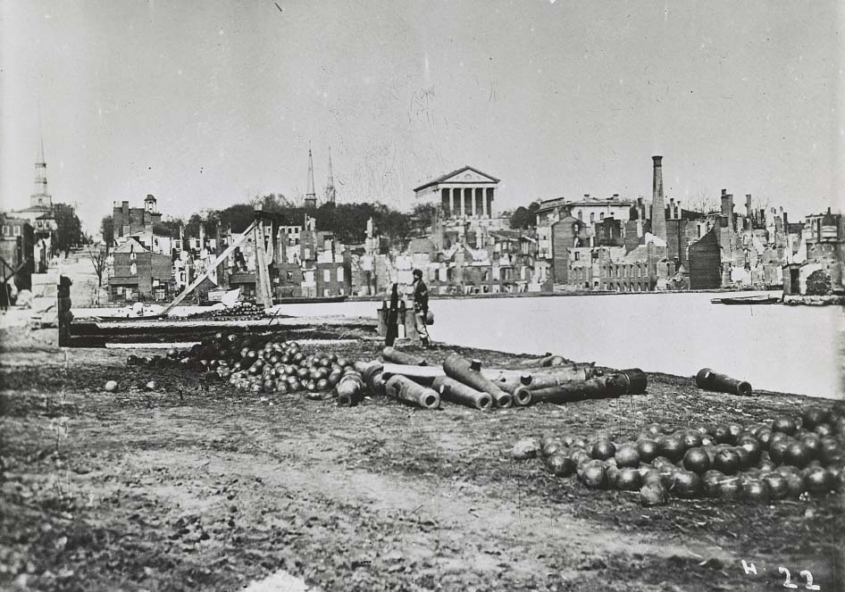 A soldier standing by a pile of cannon barrels and cannon balls with the ruins of the city in the distance, 1863