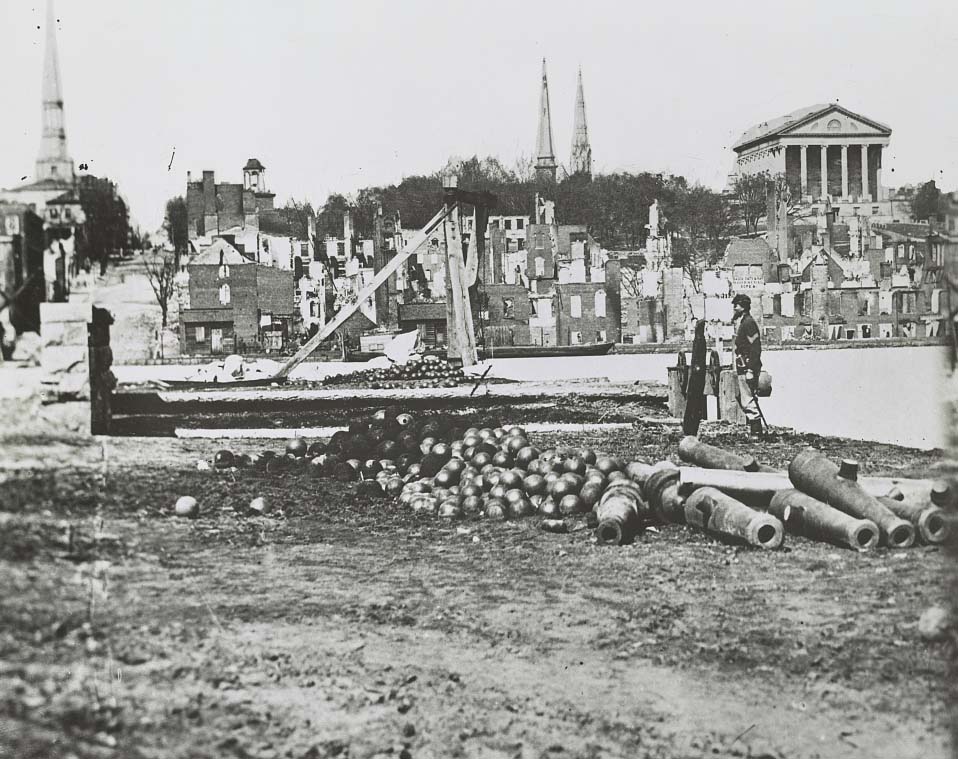 A soldier standing amid a pile of cannon balls and cannon barrels with ruins of the city in the distance, 1865