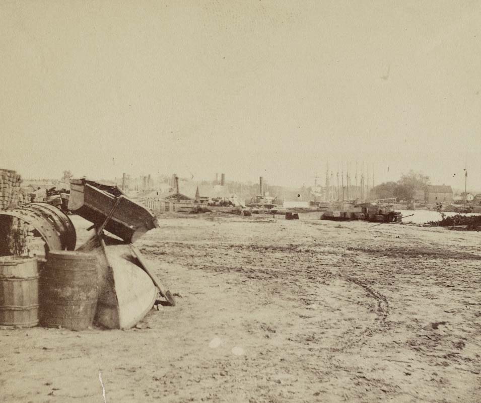 Two boats stacked next to supplies with the wharf in the distance, 1865