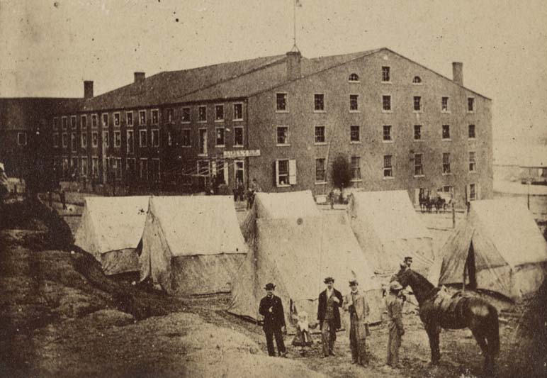 Agroup of people including a child and a man holding on to a horse next to tents in front of Libby Prison, a Confederate military prison established in 1861.