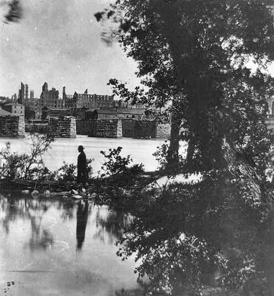 Man standing on shore overlooking ruins of bridge with ruins of city in background, Richmond, 1865