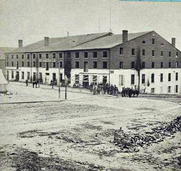 An exterior view of the site of the Confederate military prison with a group of people standing outside, 1865