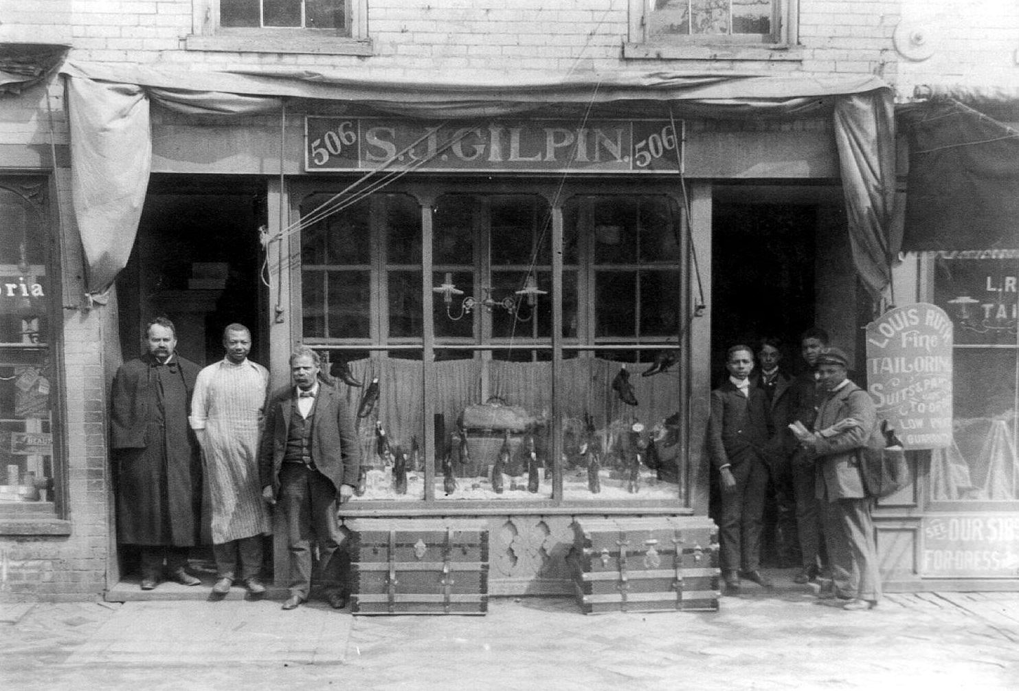 Mail carrier and six other men posed standing at entrance to shop. S.J. Gilpin shoe store, Richmond, 1900